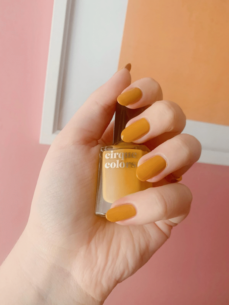 Mustard marigold short oval nails holding a bottle of yellow polish against a pink wall.
