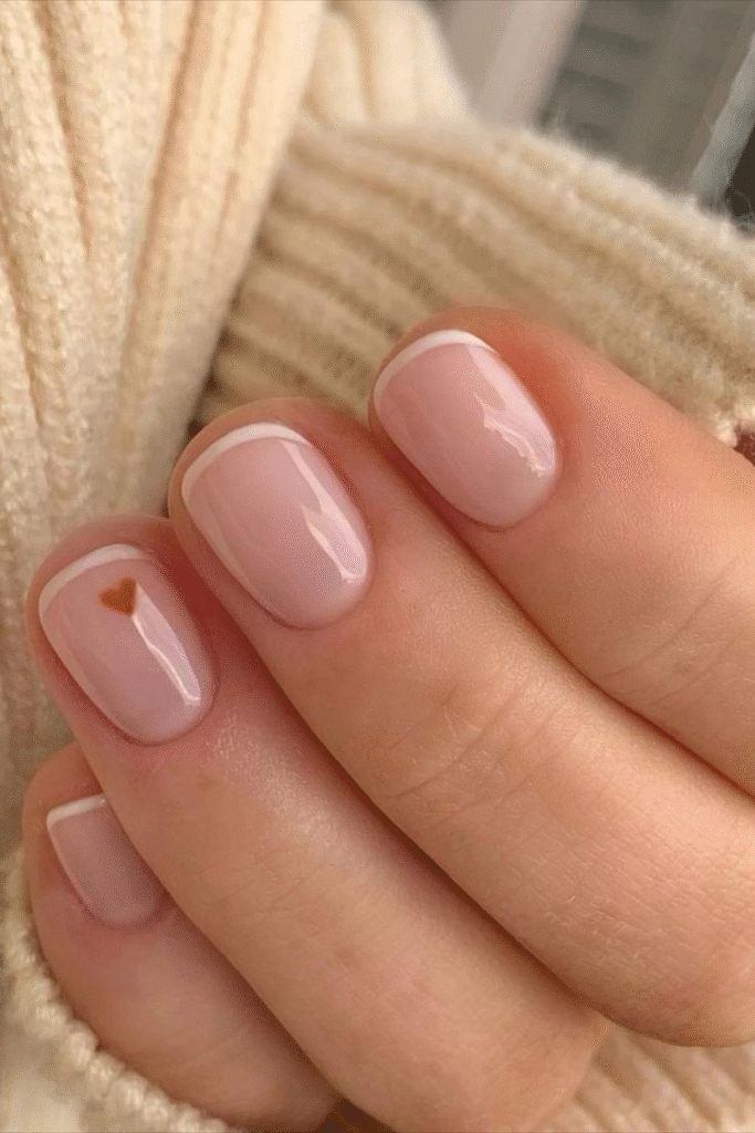 Micro French nails on short square nails with ultra-thin white tips and a tiny caramel heart accent on the pinky, over a sheer nude base.