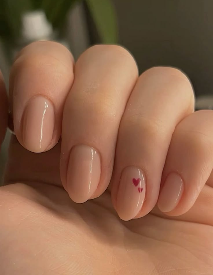 Short nude nails with glossy finish and two tiny magenta heart accents on the ring finger—minimal micro French vibe.