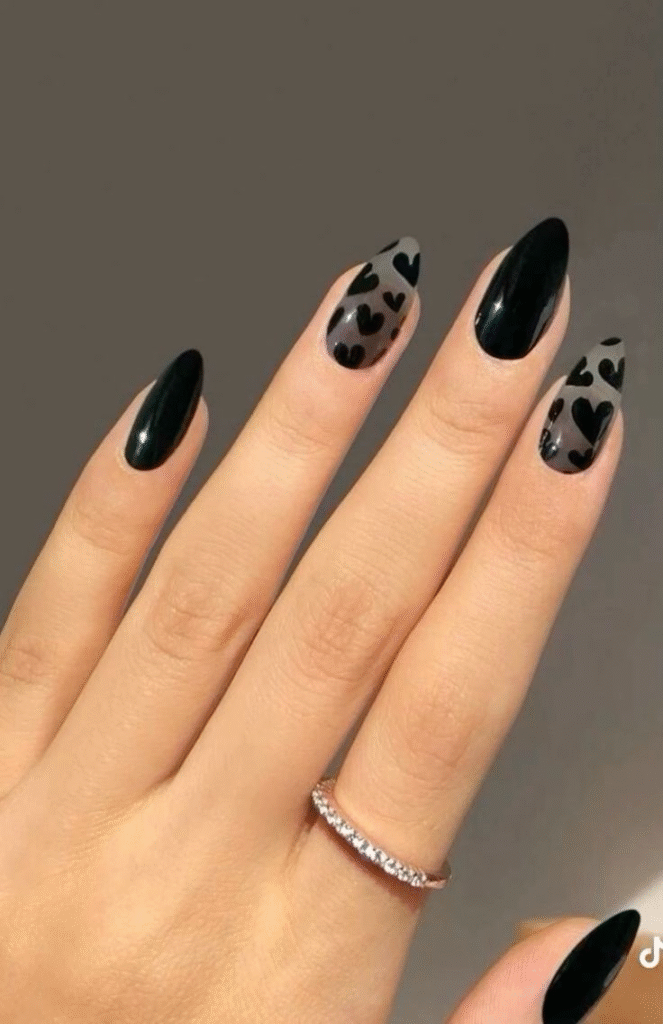 Glossy black almond nails with negative-space smoky grey hearts on two nails; delicate diamond band ring.