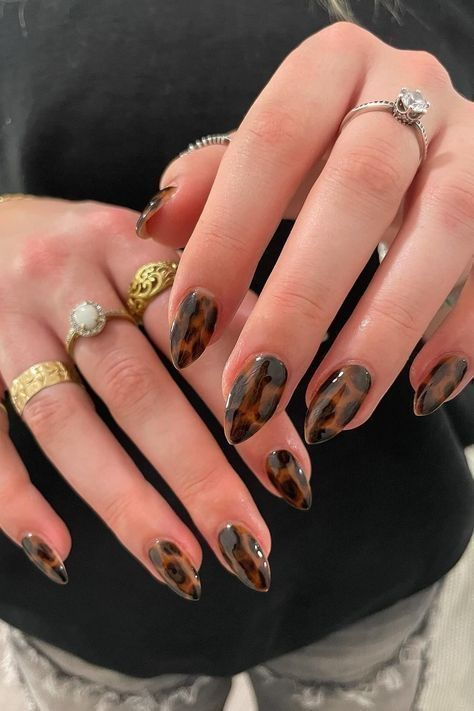 Medium almond latte tortoiseshell nails in caramel-amber-espresso jelly with a high-gloss finish; both hands posed with gold and silver rings against a black top.