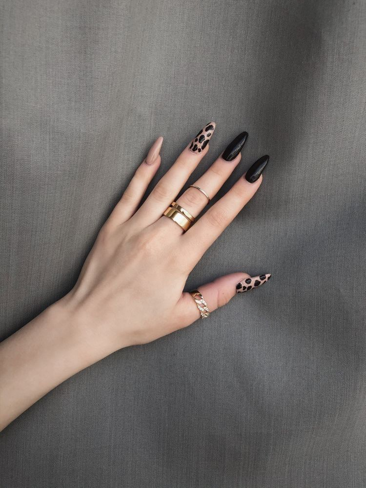 Long almond mix of glossy black solid nails and nude nails with tortoiseshell accents, gold and chain rings on a grey fabric backdrop.