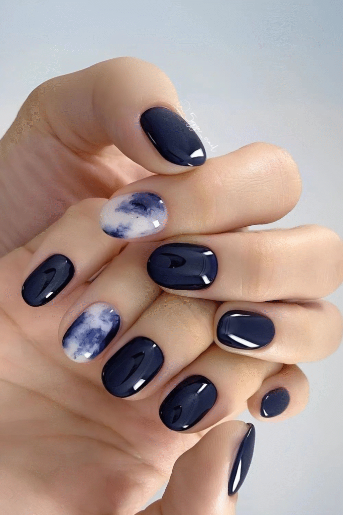 Short rounded gel nails in inky navy with one cloudy marble accent in blue and white.