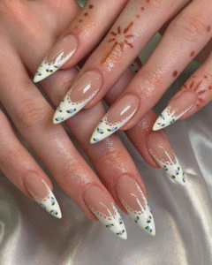 Long stiletto nails with a nude base and white French tips, scalloped lace detail, and tiny green leaf–blue bud accents; glossy finish on a satin backdrop.